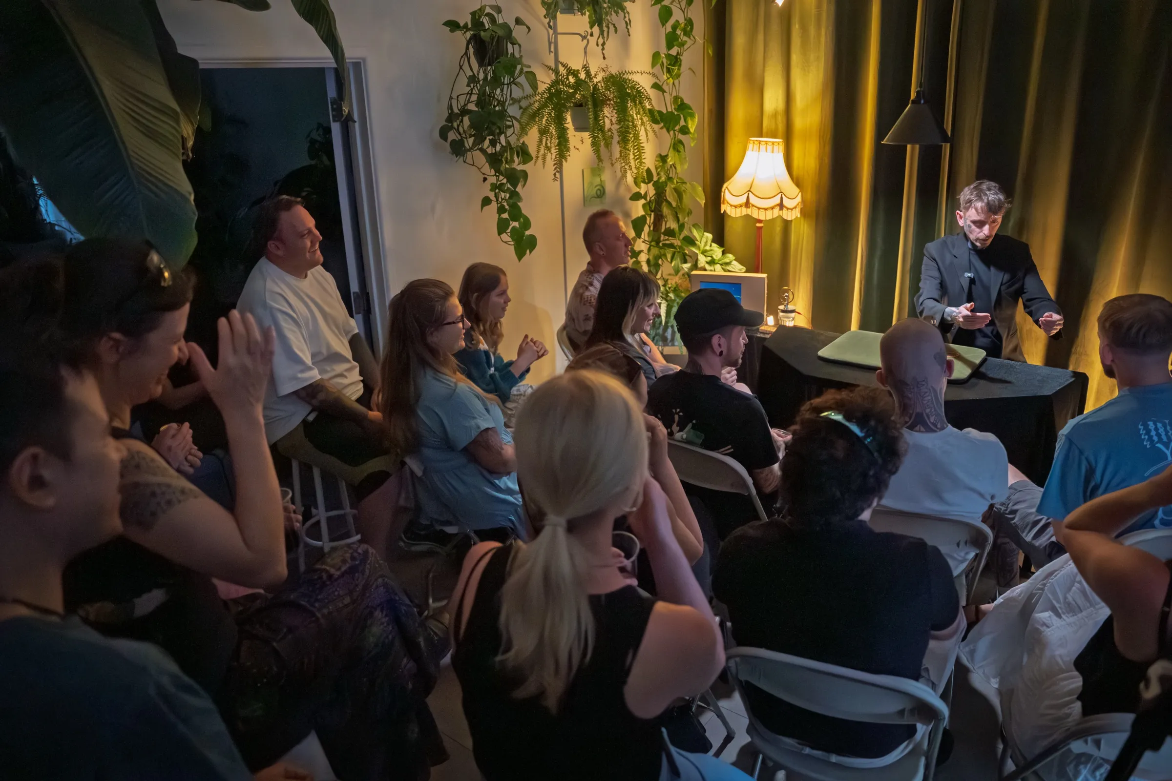 A wide view of the warm, candlelit theatre with a full audience watching the magician at the lamplit table