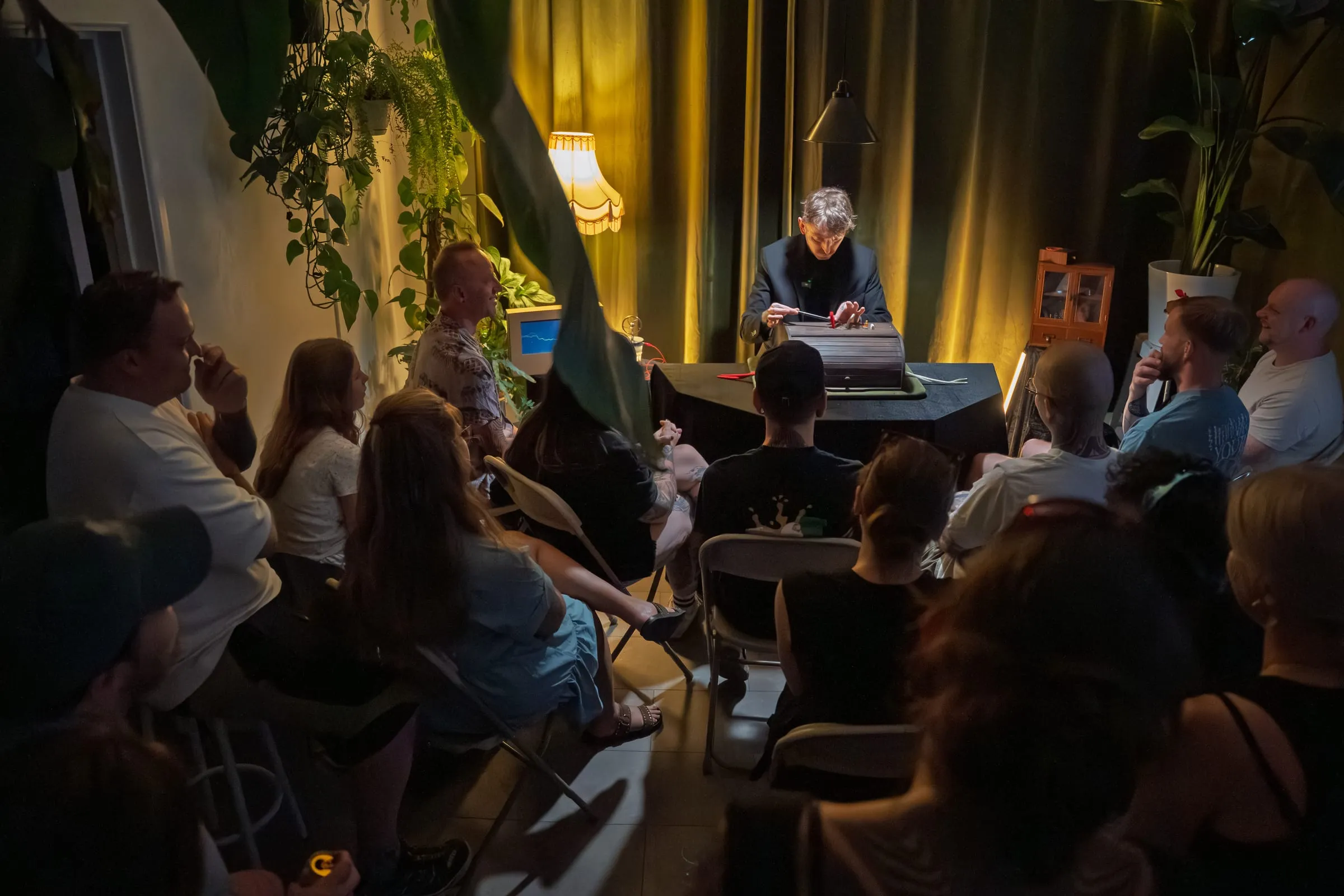 Guests seated in a half-circle of chairs around the single table, watching Si perform in the plant-filled theatre