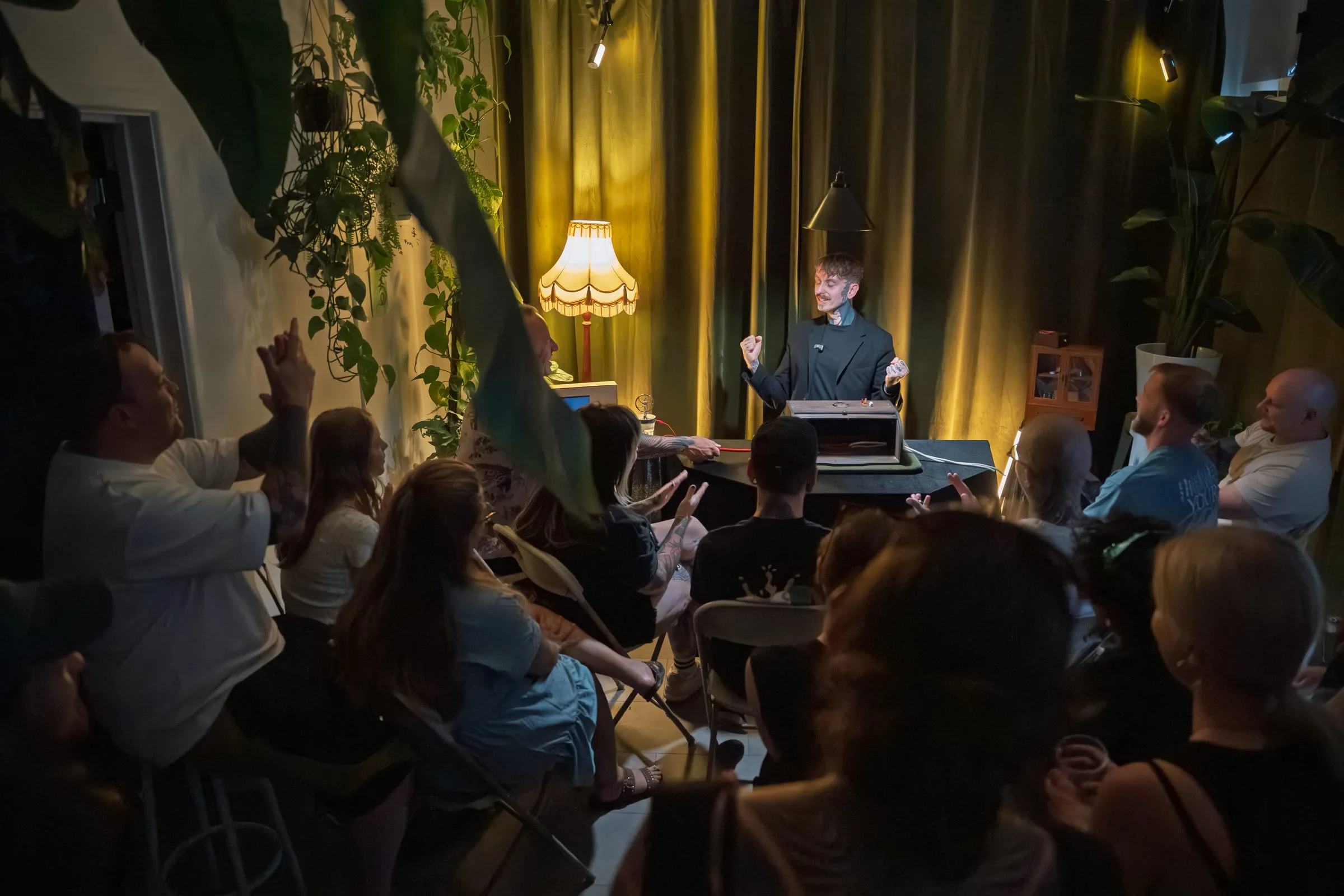 The plant-filled theatre during a show, with guests seated in a half-circle around the lamplit table